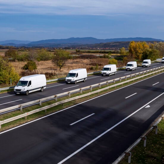 Eight white minivans are moving in a convoy on the highway. White delivery van on the highway. White modern delivery small shipment cargo courier van moving fast on motorway road to city urban suburb.